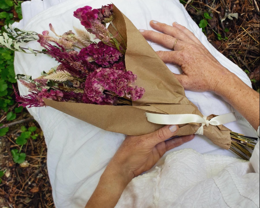A person holds a dried pink bouquet wrapped in brown paper in their lap. They are wearing a white dress. 