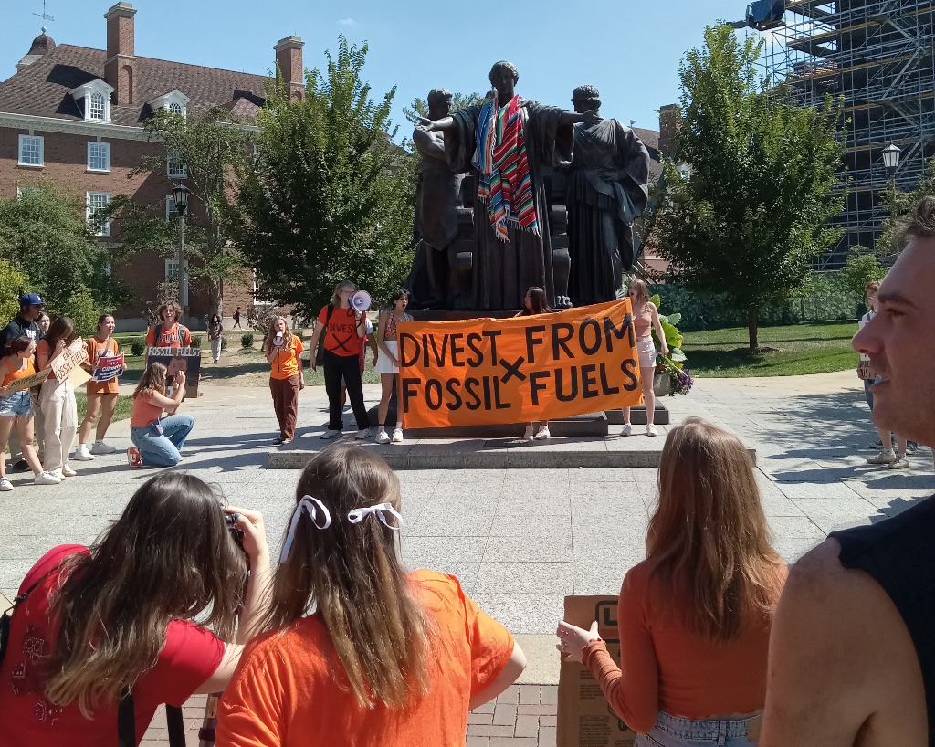 A group of students are facing the U of I Alma Mater which has a orange sign with navy writing that says "divest from fossil fuels"