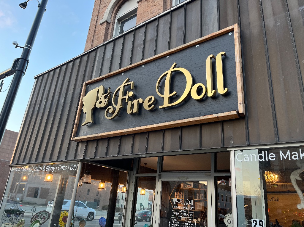 The outside of Firedoll Studio. There is a black wooden awning and a black sign with gold letters with a gold silhouette. There are glass windows below. 
