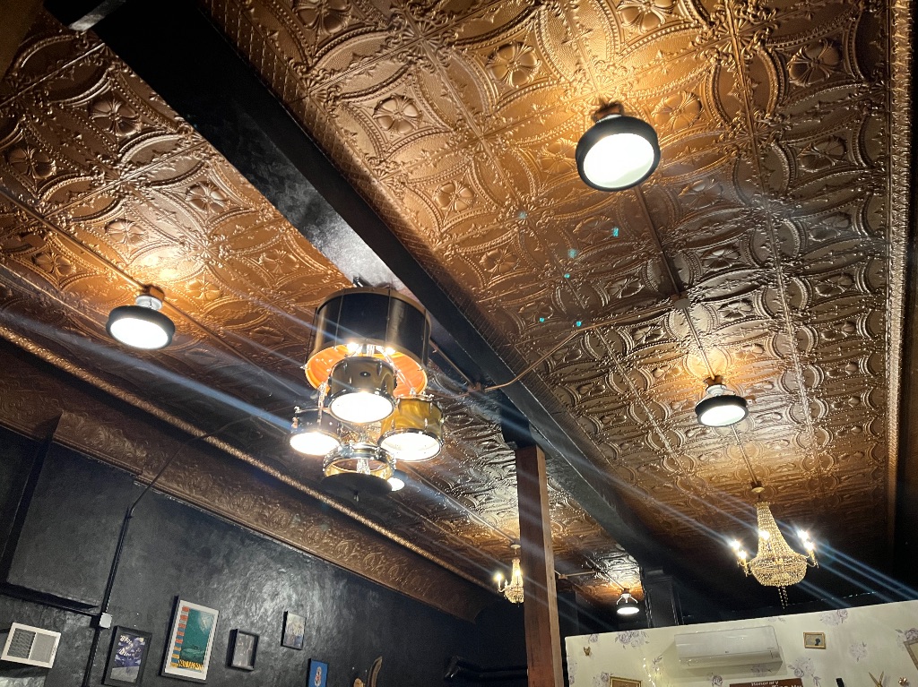 The ceiling of Firedoll. It is printed copper plates. There are lights hanging from the ceiling. In the center there is a drum made into a chandelier. 