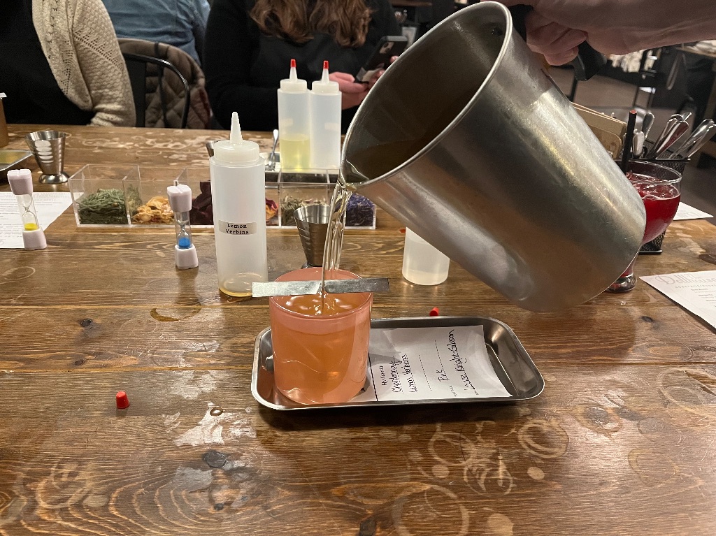 a wooden table.  A tray with a pink container with a wick and hand pouring wax from a metal pitcher. 