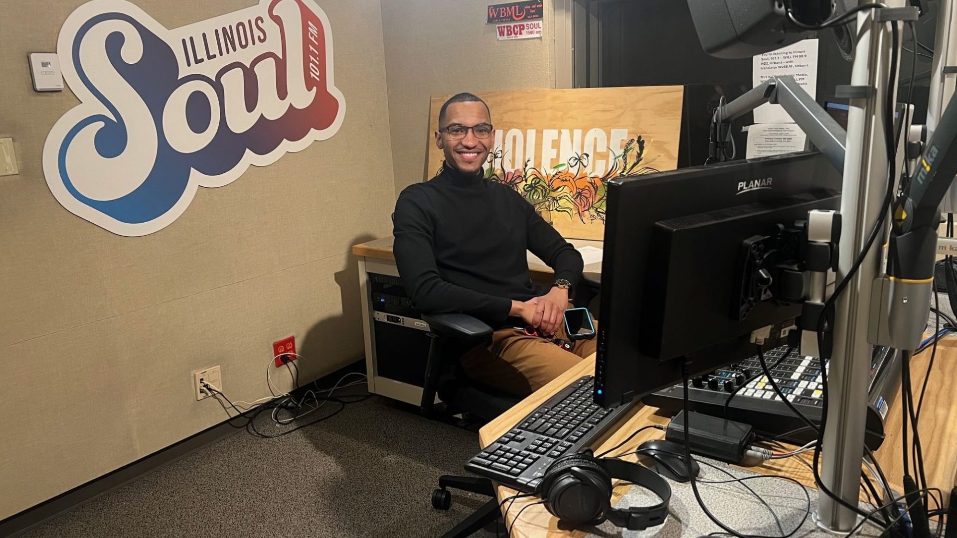 A Black man in a black long-sleeved shirt and glasses is seated at the desk in the Illinois Soul studio.