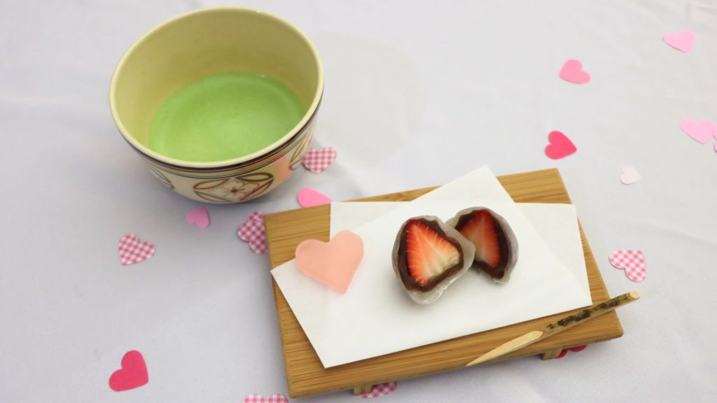 Matcha tea and a sliced strawberry coated in chocolate on a rectangular piece of white paper sprinkled with paper heart confetti.