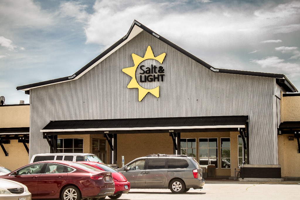 A gray building with a parking lot in front of it. The building has a salt and light logo with a yellow sun burst around the words and a cloudy sky behind the building.