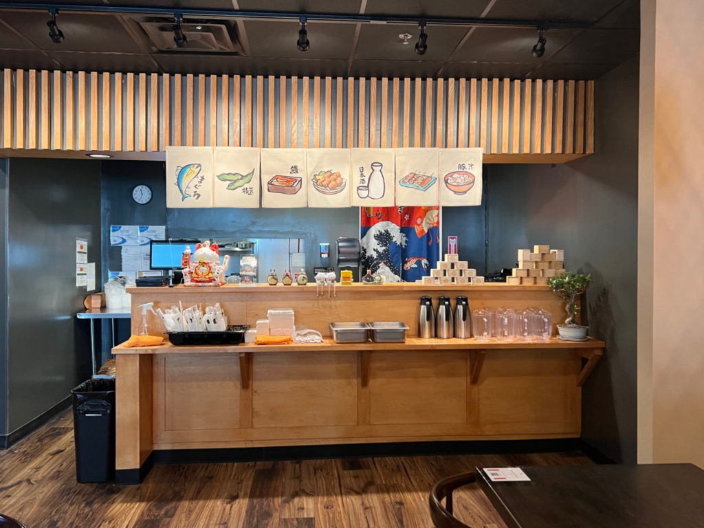 Inside Sururu Ramen, there is a sushi bar with pitchers, chopsticks, and other dining accoutrements.