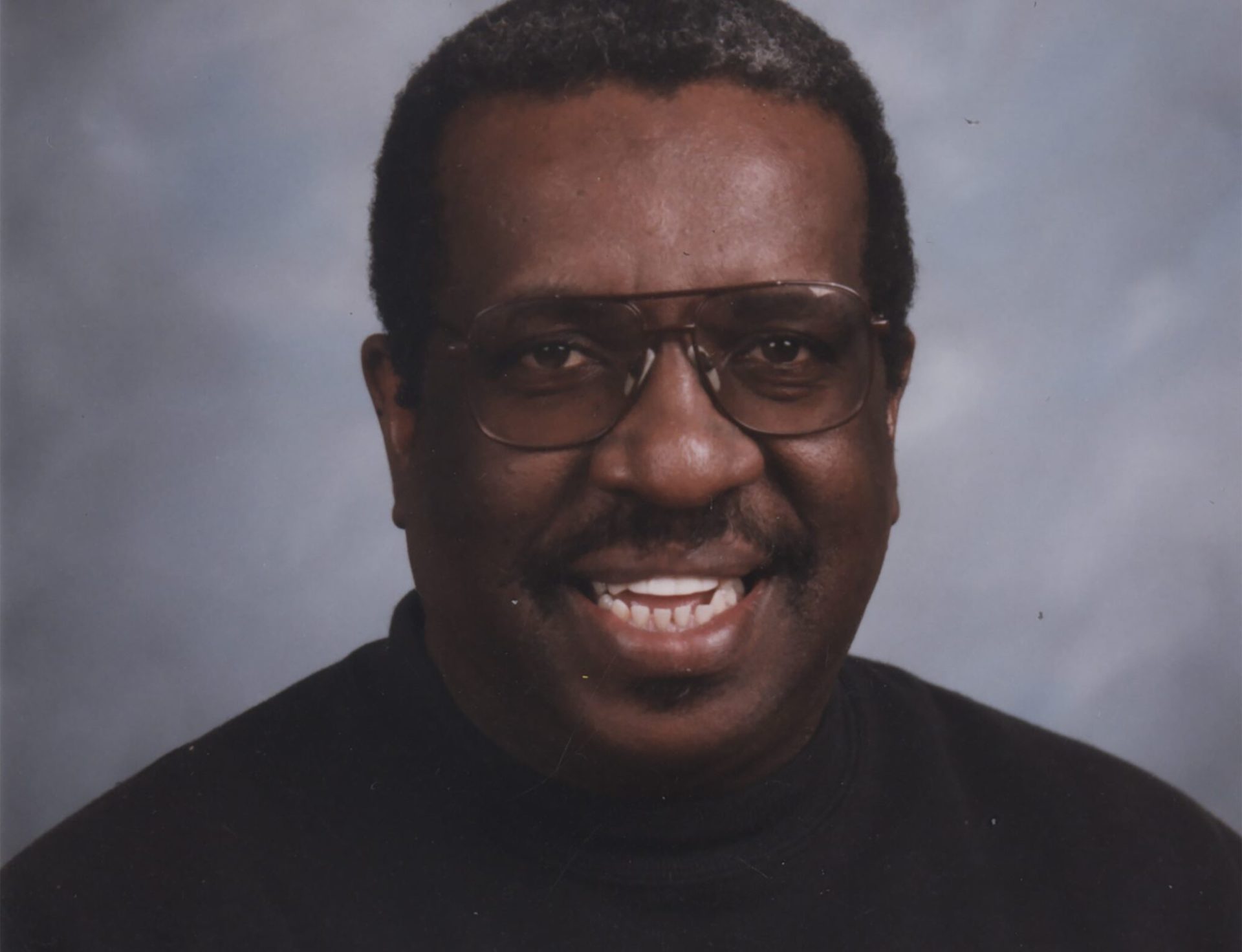 Headshot of a Black man with glasses and a black shirt. He is smiling, and has short hair and a mustache.