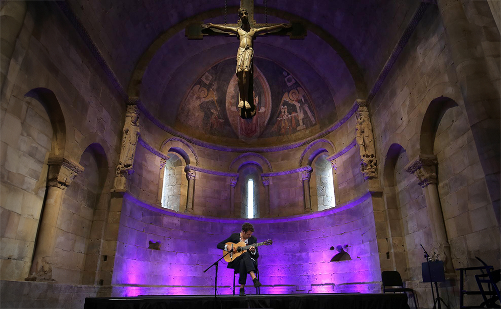 In a dimly lit historic church, a musician sits center stage bathed in a purple glow, playing an acoustic guitar. The vaulted ceilings and stone archways tower above, lit softly by ambient lights that cast warm hues on the ancient murals and carvings. To the right, musical equipment stands ready, and the crucifix sculpture above dominates the space, creating a serene yet awe-inspiring atmosphere for the performance.