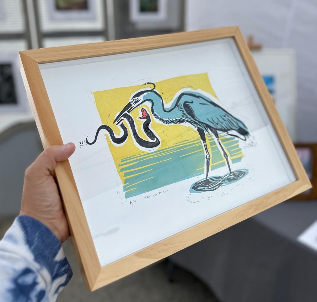 A white person is holding a print of a drawing of a heron with a snake in its mouth. The background and paper are white, the bird is blue, and the snake is black and red. The print is framed with a blond wood.