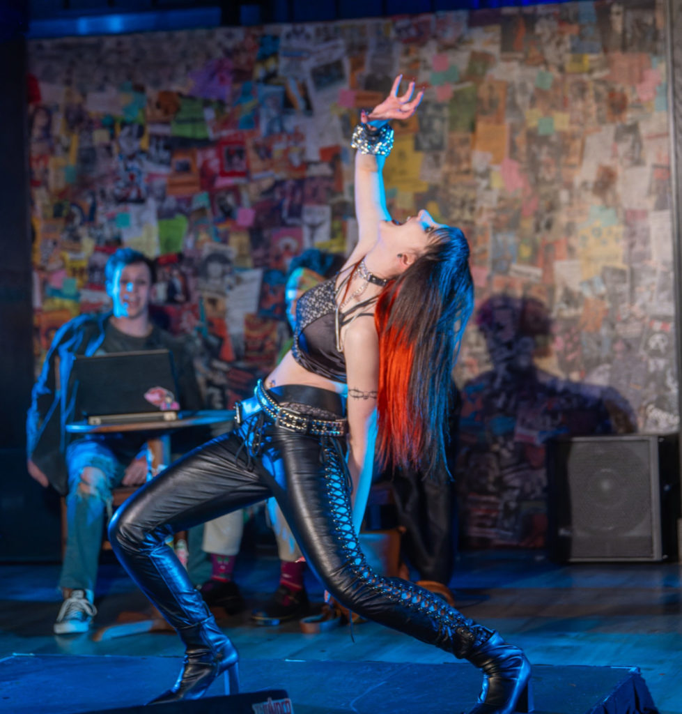 A performer is captured mid-motion on stage, with their body arched back, one arm raised high, and head tilted upwards. They have striking long hair with fiery red and orange hues on one side and deep blue on the other, and are dressed in a black, studded bralette paired with matching tight pants featuring a lace-up design on the sides. The performer is adorned with sparkling bracelets. In the blurry background, a person is sitting casually, observing, with a laptop open on a desk. Behind them is a colorful collage of various posters and images, creating a vivid and textured backdrop.