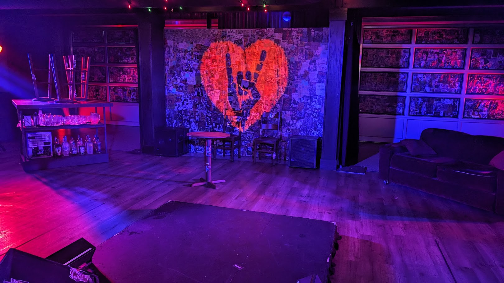 An indoor setting bathed in vibrant purple and blue stage lighting. At the center is a wall filled with a collage of posters, upon which a heart symbol with a peace sign is prominently featured. To the left, there's a bar with glasses and bottles, as well as a table with laptops. To the right, a comfortable-looking couch sits in front of a wall where framed pictures or posters are displayed. The hardwood floor reflects the colorful lighting, creating a warm ambiance. This scene appears to be from a performance space or a venue set up for an event.