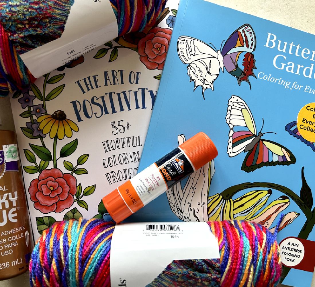 Photo of two coloring books; one is of butterflies and the other entitled the Art of Positivity. Photo also includes multicolored yarn and glue stick with orange lid.