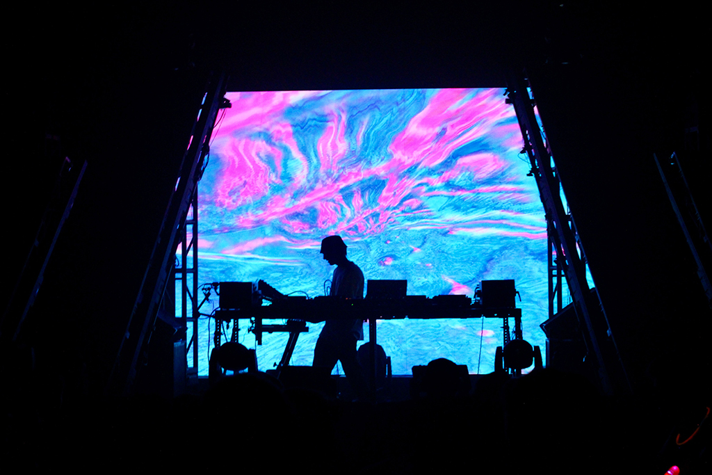 A DJ silhouetted against an intense canvas of psychedelic patterns in vivid pink and blue, suggesting a fluid, almost liquid visual experience that complements the music's rhythm and beats.