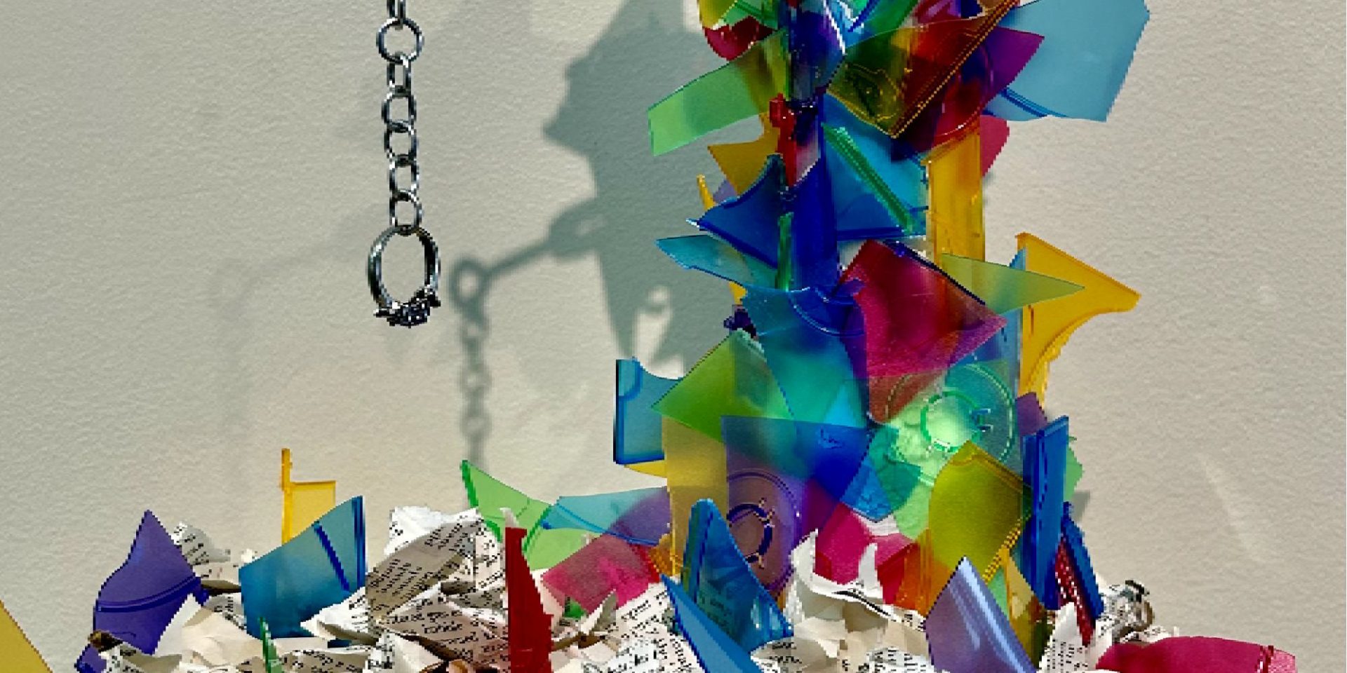Image of broken bits of colorful plastic pieces in a tower form. Crumpled and ripped book pages are at the bottom and a chain holding a ring dangles on the left, hanging from the plastic tower.