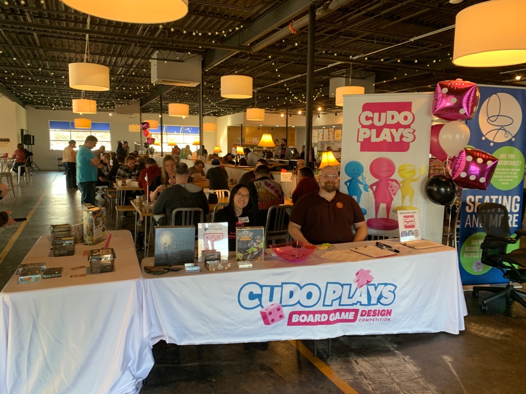 Two people are sitting at a registration table with pink dice balloons and a white CUDO Plays banner. Behind them are tables of people playing various tabletop games. 