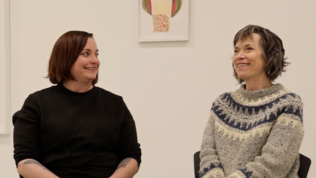 Artists Diane Christiansen and Jessie Mott, two white women, are seated in a museum space. They are viewed from the waist up, seemingly in conversation.