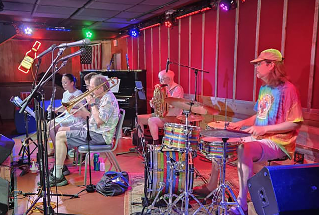 The image shows a live performance by a band with a drummer, a trombone player, and two other musicians with microphones in front of them. They are on a stage with red wall panels and blue stage lighting, and there's an American flag hanging in the background. The band members are casually dressed, with the drummer wearing a tie-dye t-shirt and hat, reflecting a laid-back, possibly jazz or folk, ambiance.