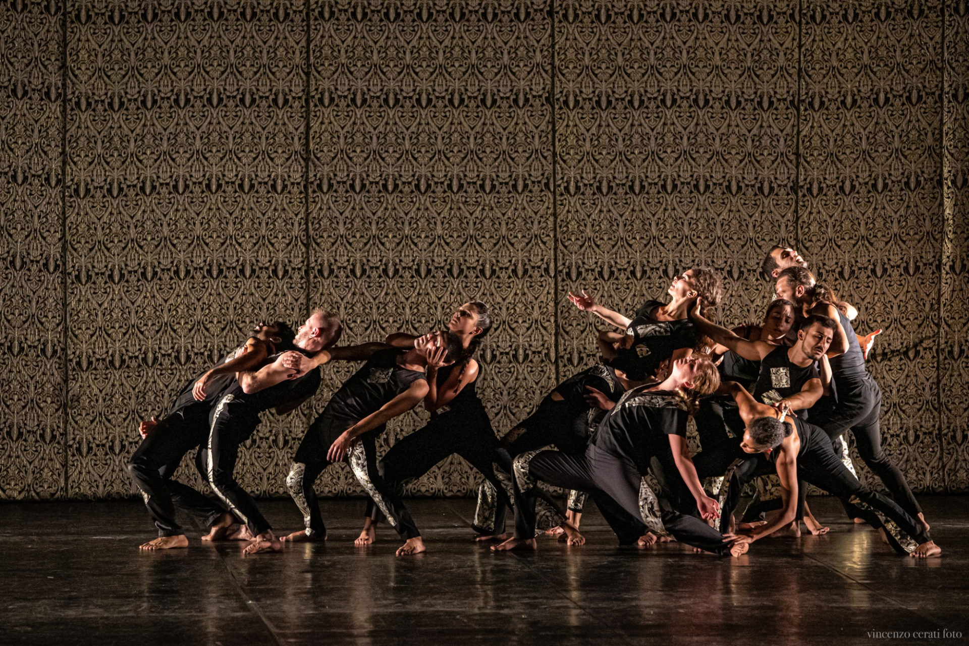 Approximately 12 people dancing on a dark stage, with a highly decorative black and gold backdrop. The dancers are men and women, wearing black outfits with gold details. They are in a group where some people are bending and leaning backward onto each other.