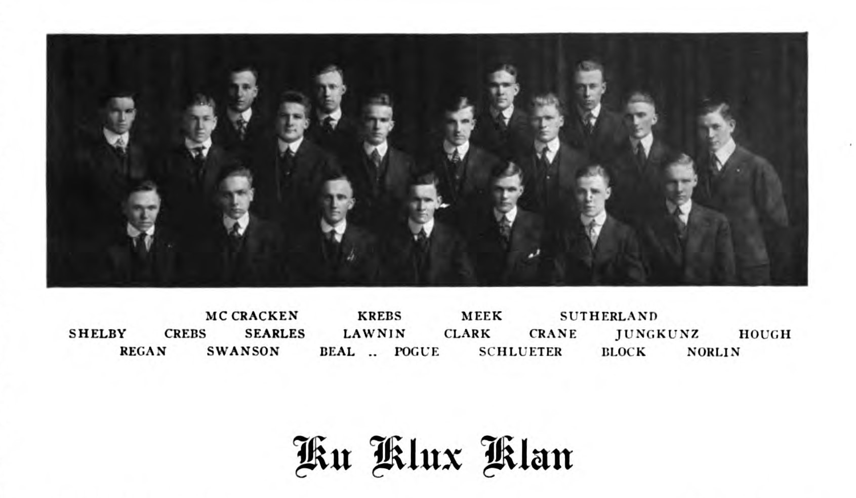 A black and white photo from the Illio yearbook of student KKK members. The group of white men are sitting and standing in straight lines wearing black suits with white shirts in front of a dark curtain. The members are listed below the picture.
