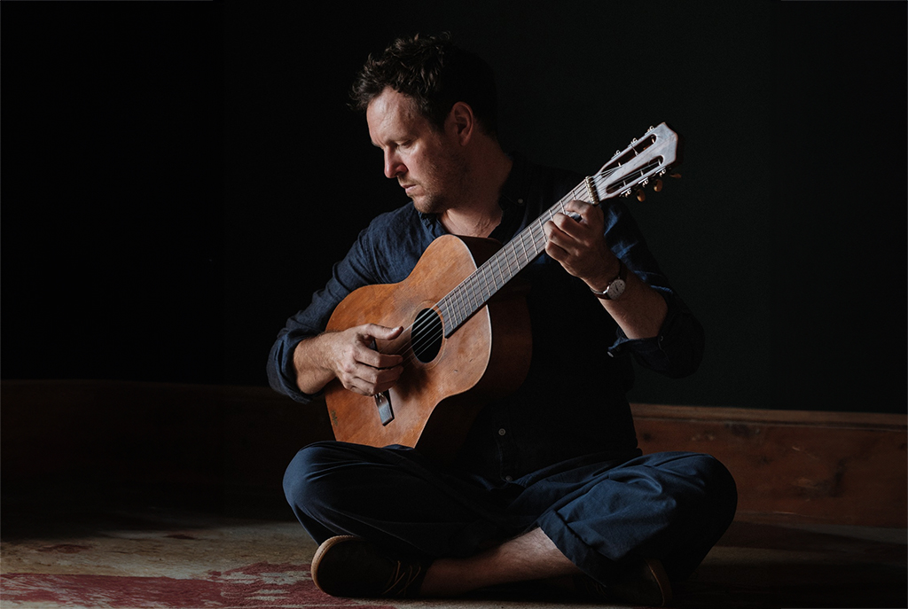 Seated cross-legged on a textured rug, a musician holds a classical guitar in a dimly lit, intimate space. His casual, yet thoughtful attire contrasts with the solemnity of his pose and expression, which reflects a deep concentration on the music. The natural light from above casts soft shadows, enhancing the contemplative mood of the setting.