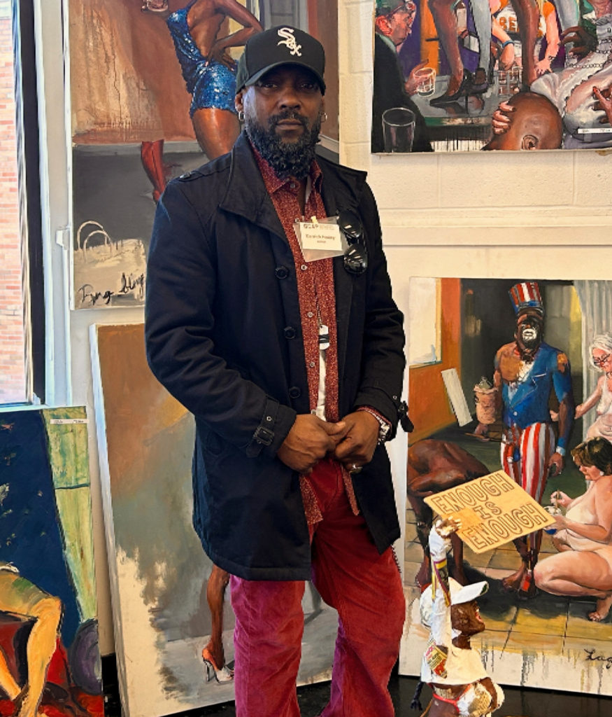 Photo of a Black man wearing a dark blue overcoat and a Chicago White Sox ball cap. He is standing in front of his paintings of images of Black and white figures in a variety of poses.