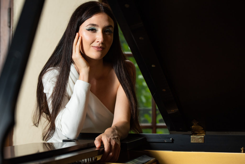 A women with long dark hair and a white off the shoulder top, leaning on a black grand piano.
