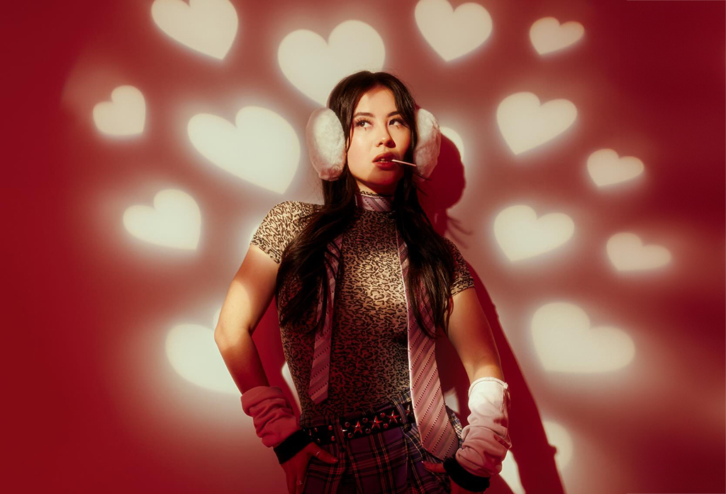 an individual against a red background adorned with soft-focused heart shapes. They wear fluffy white earmuffs, a patterned turtleneck, a plaid skirt, and white gloves, striking a confident pose with hands on their hips.