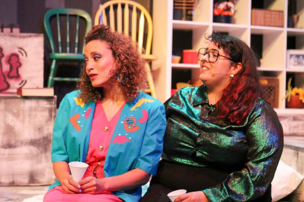 Photo of two women on a theatre set. The one on the left has curly hair and is wearing a pink dress with a teal jacket. The woman on the right is wearing a sparkling green shirt and wears glasses.