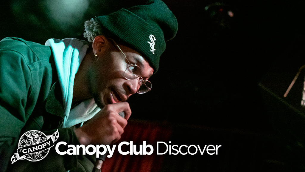 The image features a male performer with a focused expression, wearing a green baseball cap, clear-framed glasses, and a light blue jacket with white accents. He is holding a microphone close to his mouth, suggesting he is either singing or speaking. In the background, the setting appears to be dimly lit, characteristic of a club or concert venue atmosphere. The image is branded with the Canopy Club logo and the text "Canopy Club Discover," indicating that this may be a promotional image for a discovery series event at the venue, highlighting new or emerging artists.