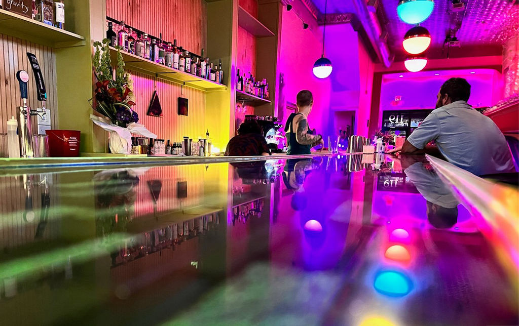 In a lively bar aglow with neon reflections, patrons enjoy a relaxed ambiance. Vivid pink and yellow hues illuminate the backdrop, accentuating a modern bar stocked with an array of bottles. The bar's surface gleams, mirroring the colorful overhead lights that dangle like illuminated orbs in a spectrum of reds, blues, and greens. A bartender, engaged in the art of mixology, attends to the guests' requests, while a patron sits contemplatively, perhaps pondering the day or simply soaking in the vivid, convivial atmosphere. The scene is a tapestry of color and light, inviting and electric, promising a warm welcome to all who enter.