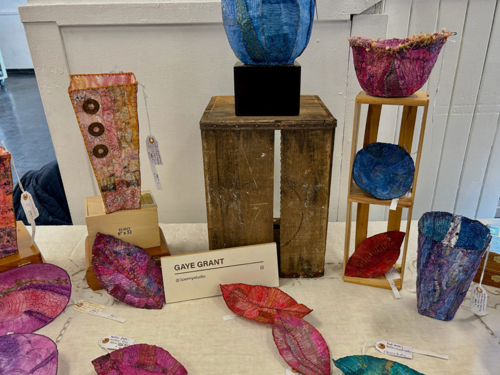 Photo of pink, purple, and blue textile art in the shape of vases and leaves. Gaye Grant @3pennystudio is on the tag in the middle of the table.