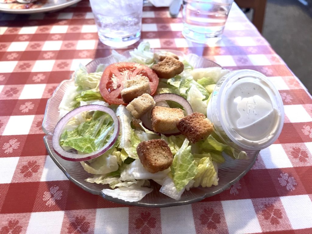 A garden salad on a clear plate with lettuce, croutons, tomatoes, and red onion, with a small plastic container of ranch dressing,