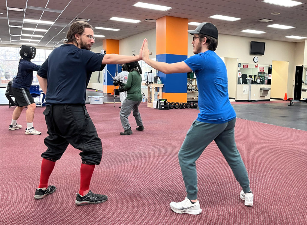 Two white men in athletic clothing practice historical fencing. They are facing each other with their left hand (right) and the right hand (left) pressed palm to palm. There are other fencers in the background practicing.