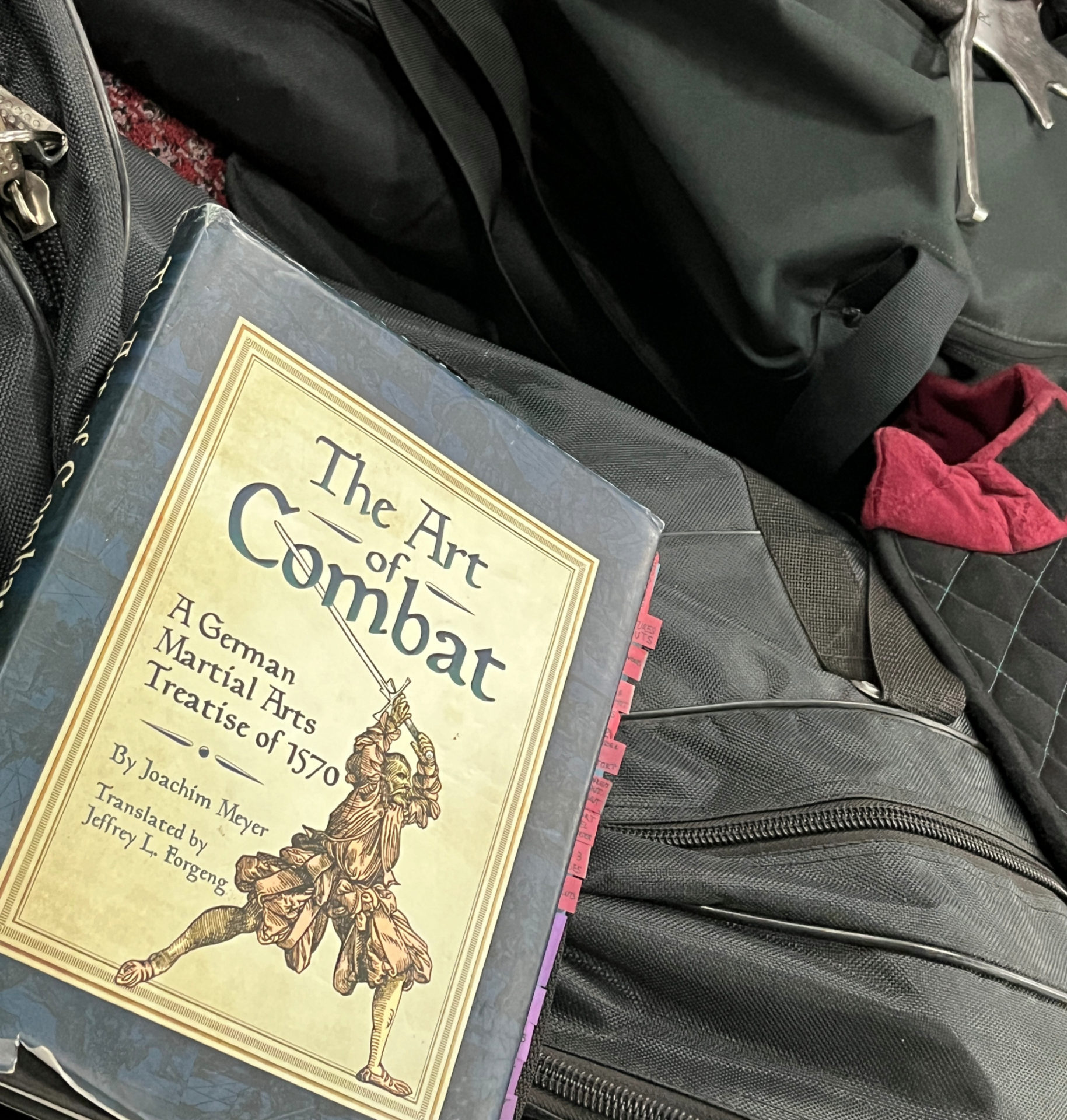 The Art of Combat by Joachim Meyer is sitting on a pile of athletic and protective gear. The book contains many tabs on the side, where the reader made notes.