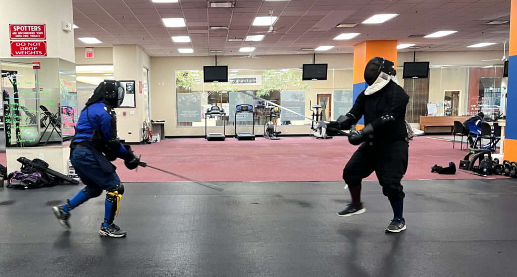 Two men in fencing protective gear square off with swords in a gym. They are standing across from each other with their swords pointed toward each other.