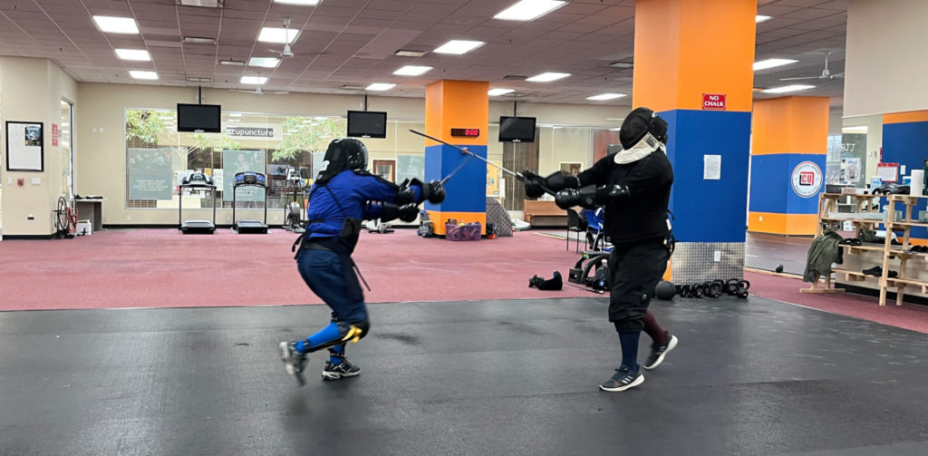 Two men in fencing protective gear square off with swords in a gym. They are standing close to each other, and their swords are crossed in between their bodies.