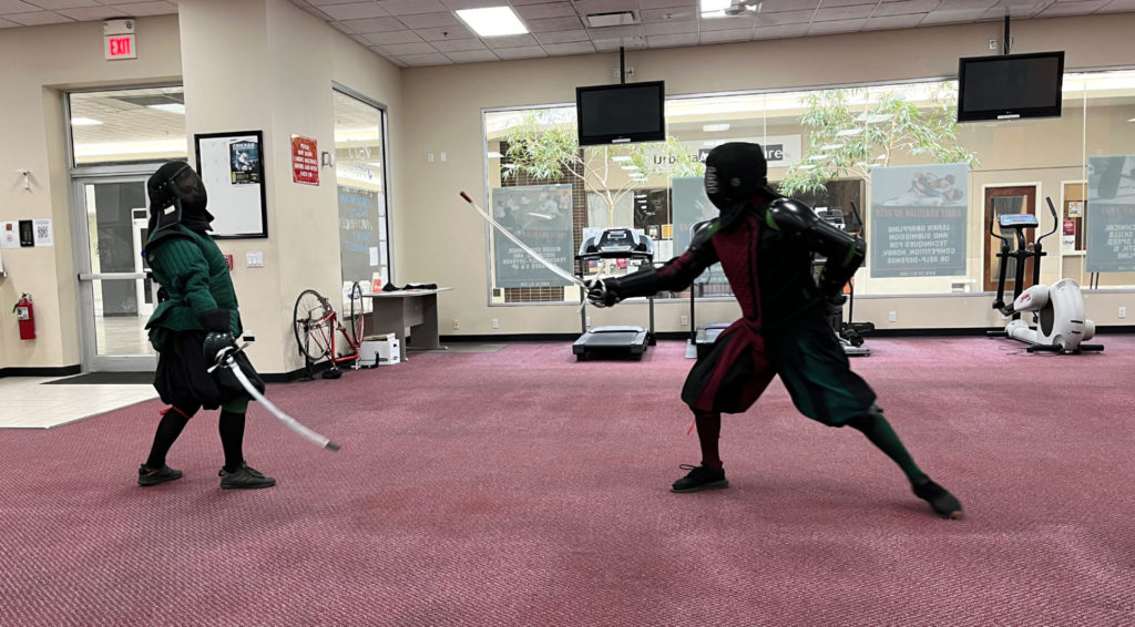 Two men in fencing protective gear square off with swords in a gym with a faded red carpeted floor. They are standing across from each other. The man on the left wears green and is leaning away as the man in the right (in red) lunges forward with his sword pointed in the green man's direction.