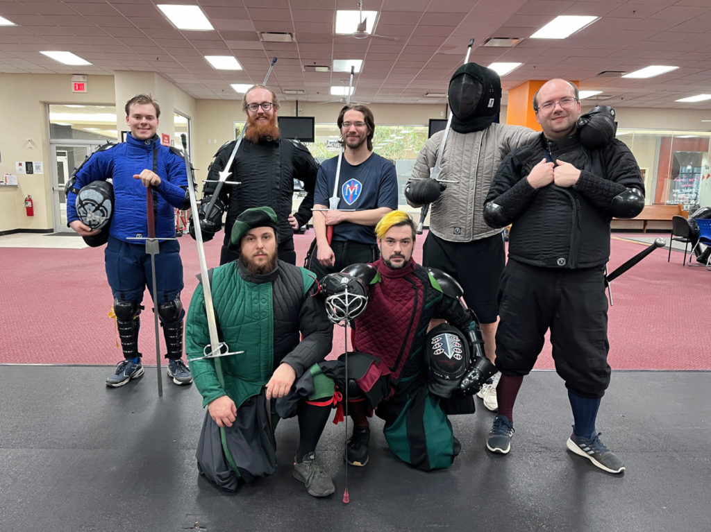Seven white men, one in a fencing face mask, pose after a Tempered Mettle practice session. The five men in the back row are standing, and the two men in the front row are kneeling. They are in a gym. Most of them are holding longswords.