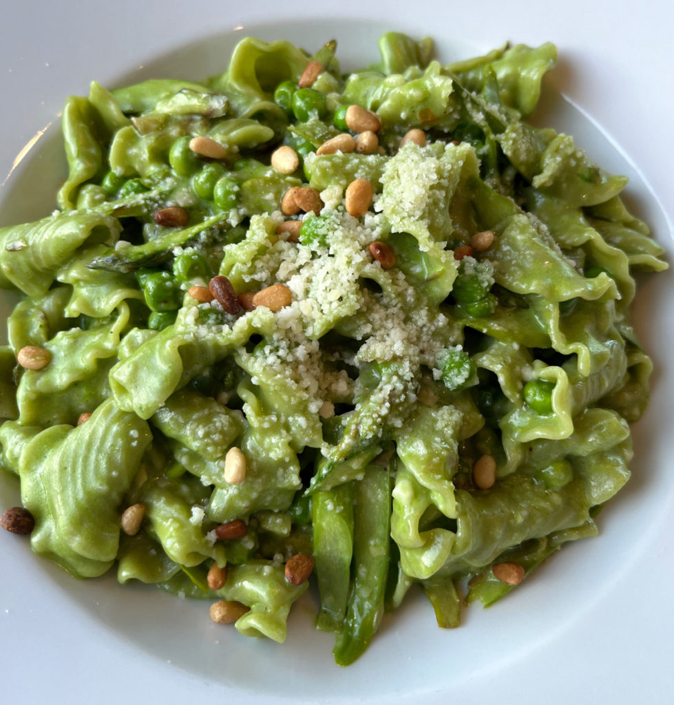 Green bell-shaped pasta served in a white bowl with shaved asparagus, peas, and pine nuts and a sprinkling of Parmesan cheese.