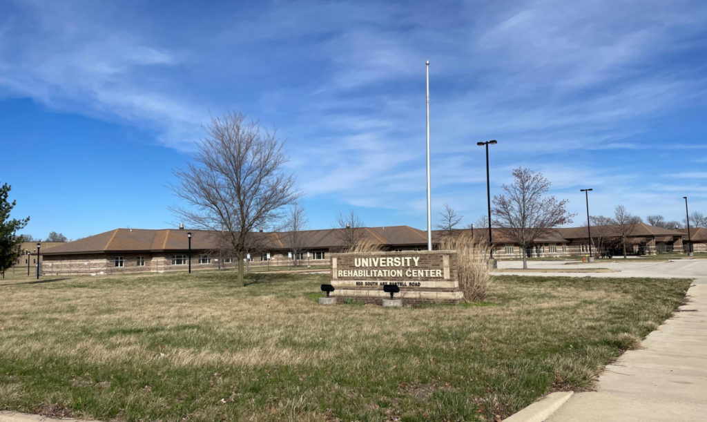 The Champaign County Nursing Home / University Rehabilitation Center in Urbana. A sign is out front, behind which is a large, single story building that is closed. The sky is very blue with wispy clouds.