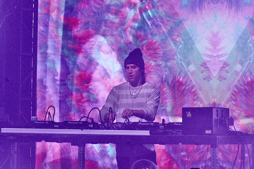 A DJ performing on stage, positioned behind a DJ console with various equipment. They are wearing a striped long-sleeve shirt and a beanie, giving off a focused vibe as they work the decks. The backdrop is a vibrant mix of colors and patterns that create an abstract, tie-dye effect, enhancing the visual atmosphere of the performance.