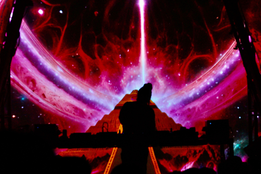 A DJ is silhouetted against a cosmic and galactic backdrop, with hues of pink and purple and a beam of white light emanating from the center, giving the impression of interstellar travel or a portal to another dimension.