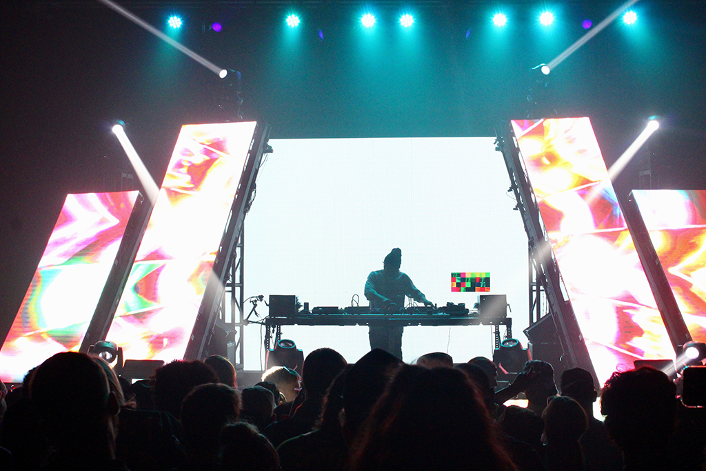 A DJ booth is silhouetted against a massive screen filled with kaleidoscopic, colorful imagery. Bright stage lights at various angles create a dynamic lighting effect, while the crowd in the foreground appears as darkened figures, their attention directed towards the performer, whose silhouette is the focal point of the composition.