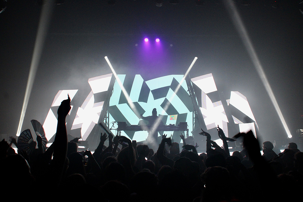 A DJ performing before a lively crowd, with hands raised in the air. The backdrop consists of an intricate, geometric black and white design that forms a stark, visually arresting contrast with the silhouetted figure of the DJ, suggesting a high-energy climax to the event.