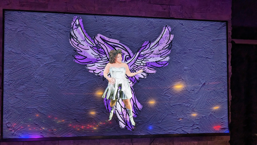 Displayed on a large screen is a digitally added image of a person with stylized white wings. The person is wearing a shiny silver dress and is posed with one arm extended outward, holding a microphone. The background of the screen has a textured appearance, predominantly in purple hues, with small lights scattered across the bottom creating a subtle, glowing effect.
