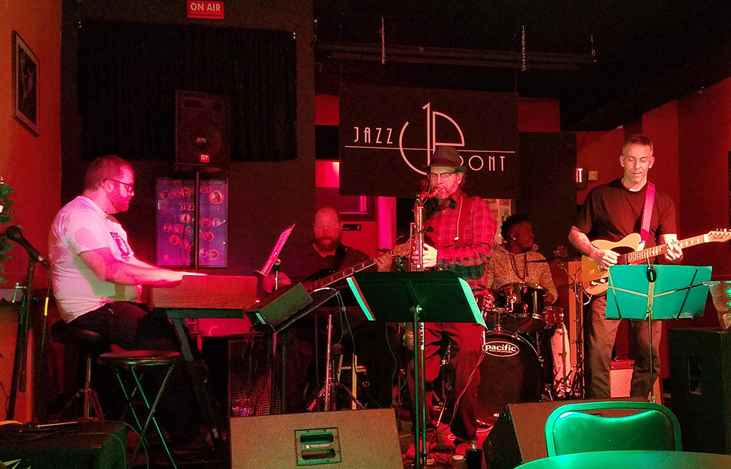 A live jazz band performance, with a keyboardist, drummer, saxophonist, and guitarist, bathed in warm stage lighting with a "Jazz Up Front" sign in the background.