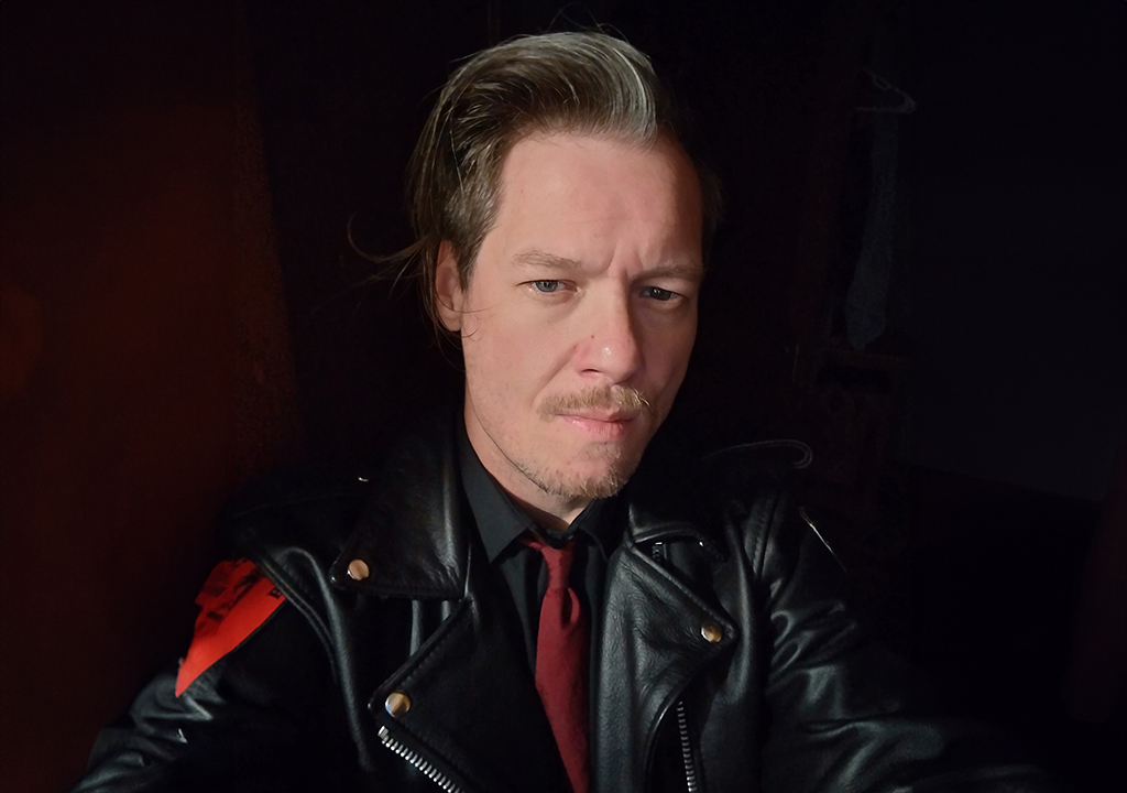 A person with a contemplative expression, wearing a black leather jacket with a red armband on the right sleeve. The background is dark, which emphasizes the individual's light complexion and the details of the leather jacket.