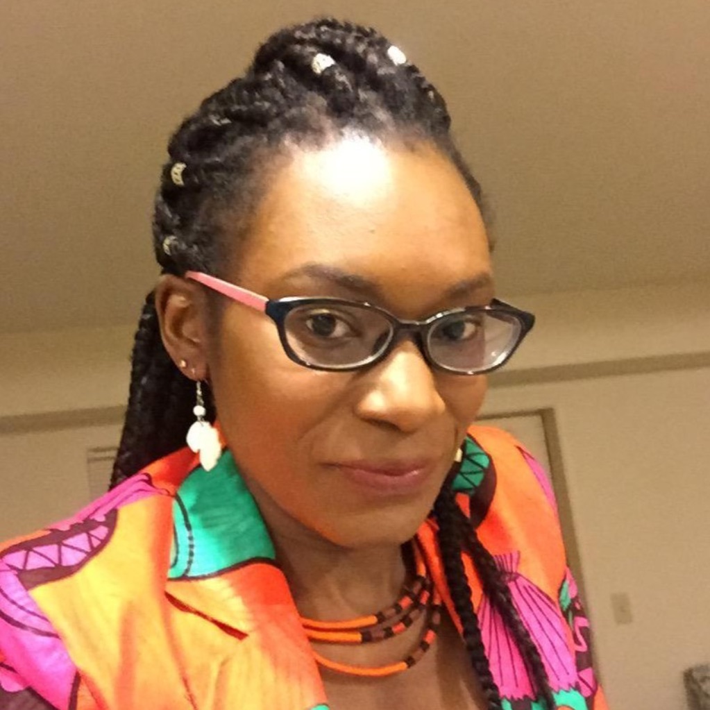 A black woman stares at the camera, she is wearing a colorful jacket of orange, pink and green, and black glasses and earrings. 