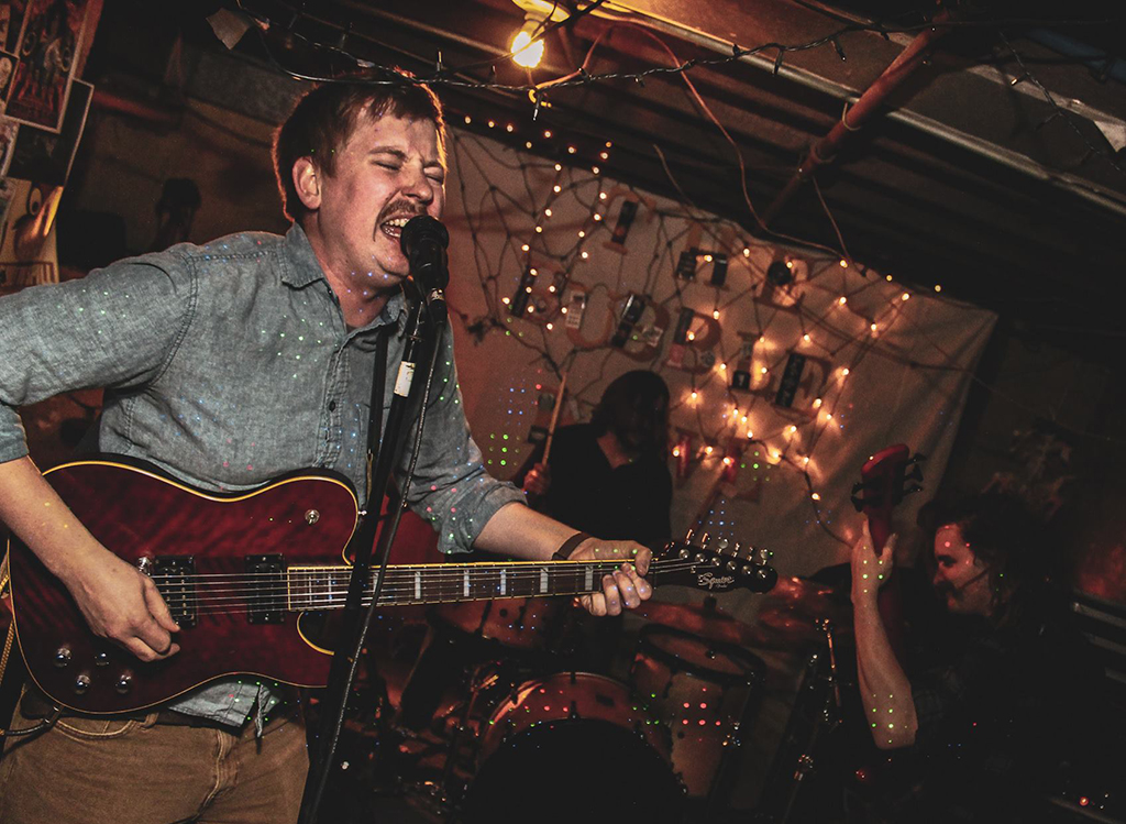 A musician in a blue shirt, passionately singing and playing a red electric guitar, bathed in the warm, ambient light of string lights in what appears to be a cozy, indoor venue. The energy and emotion of the performance are palpable, with the musician's expression conveying intensity and fervor. You see a drummer and a bass player in the background playing with equal passion. The surroundings suggest an intimate concert setting, with a casual and engaged atmosphere.