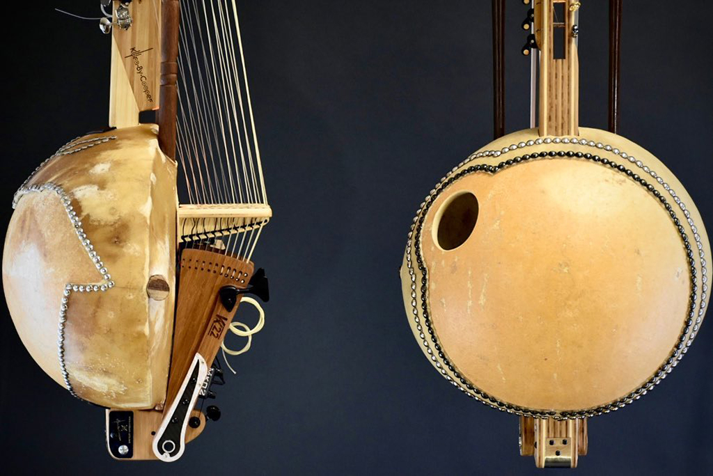 Two traditional African stringed instruments, known as koras, hang against a dark background. Their large, rounded bodies are similar to that of a lute, embellished with what appears to be tacks or studs. The necks of the instruments are long and feature numerous vertical strings, which suggests a complex sound and playing technique. The craftsmanship highlights the cultural heritage and musical traditions these instruments represent.