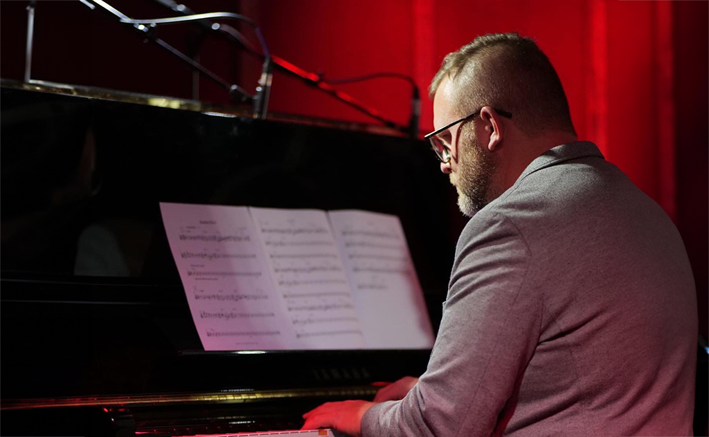 The image shows a male pianist playing a black piano. He is viewed from the side and is focused on sheet music in front of him. The pianist is wearing glasses and a gray jacket, and the background is a deep red, which gives the setting an intimate and warm feel, typical for a jazz club or a concert venue with a cozy ambiance. The presence of sheet music suggests that he is either practicing, performing, or accompanying another musician or group.